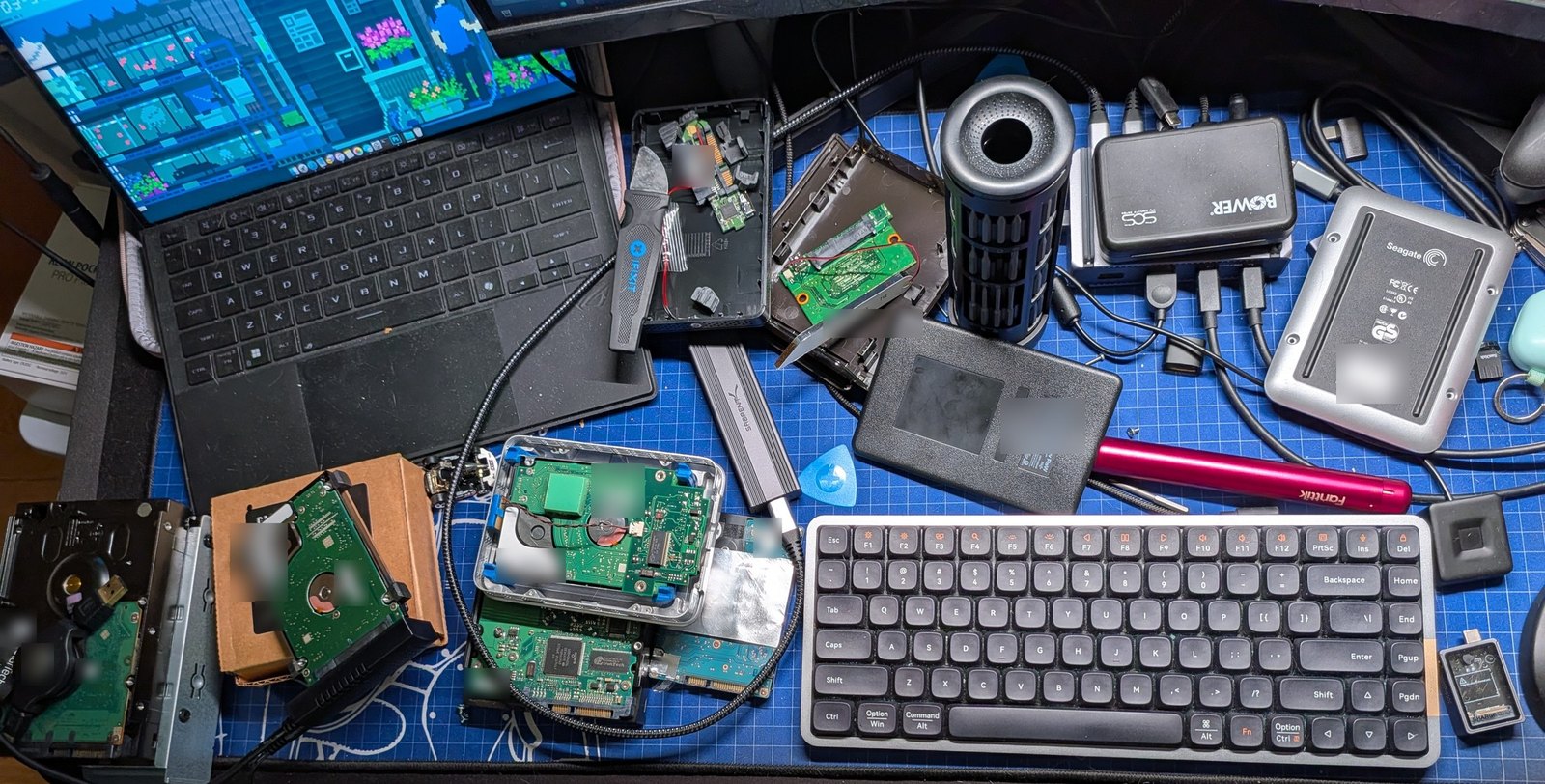 A workbench covered with bare hard drives, USB enclosures and adapters, with two laptops running recovery jobs